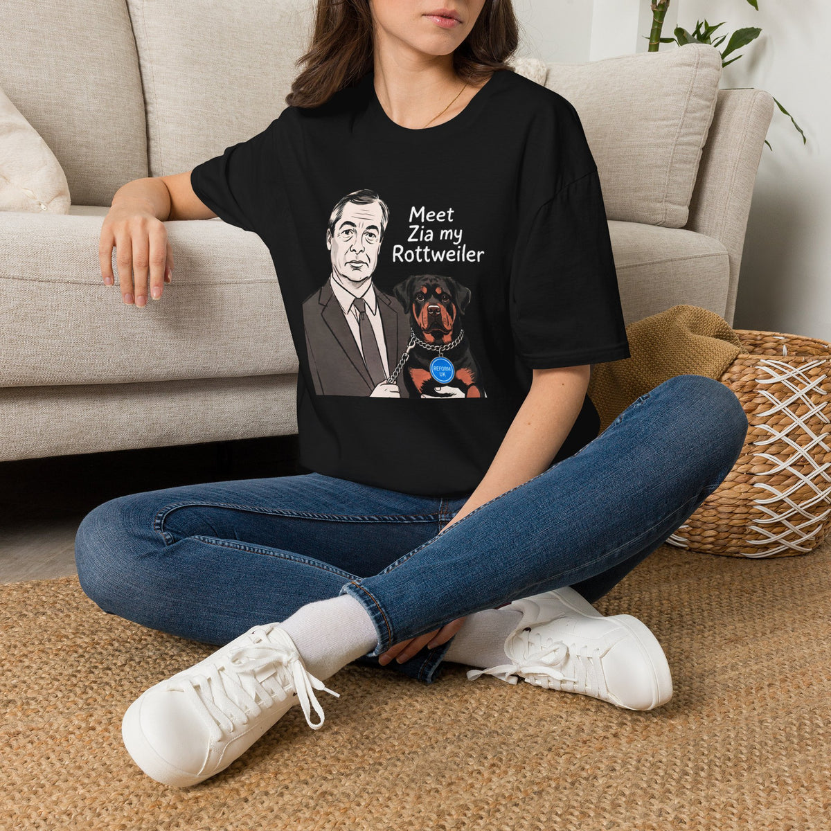 A woman sitting cross-legged on a woven rug in front of a beige sofa, wearing an oversized black graphic T-shirt with a portrait of NIGEL FARAGE  in a suit and the text 'Meet Zia my Rottweiler'.