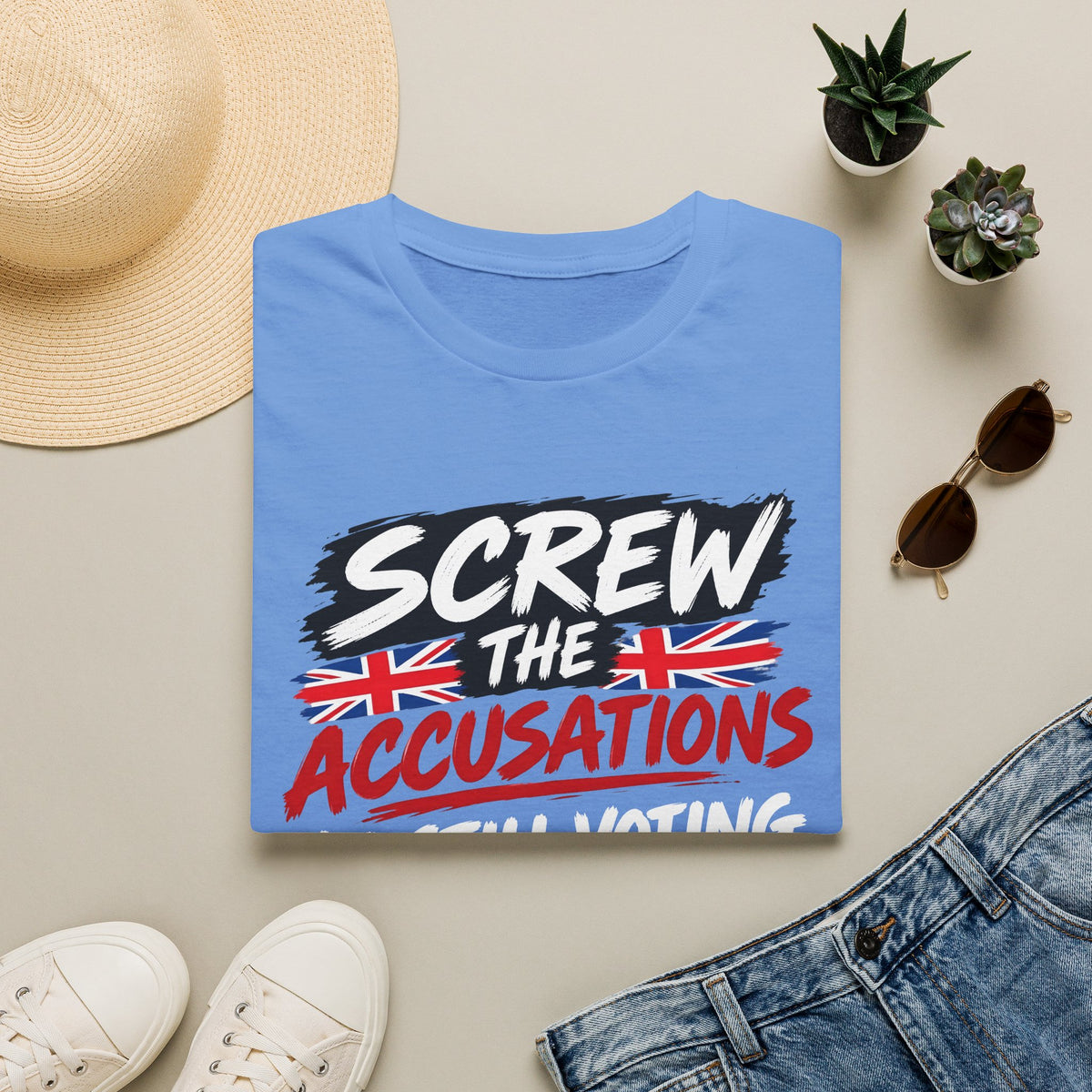 A folded light blue graphic T-shirt is arranged in a styled flat lay on a light beige surface. The shirt features the text 'SCREW THE ACCUSATIONS' with Union Jack flag graphics.