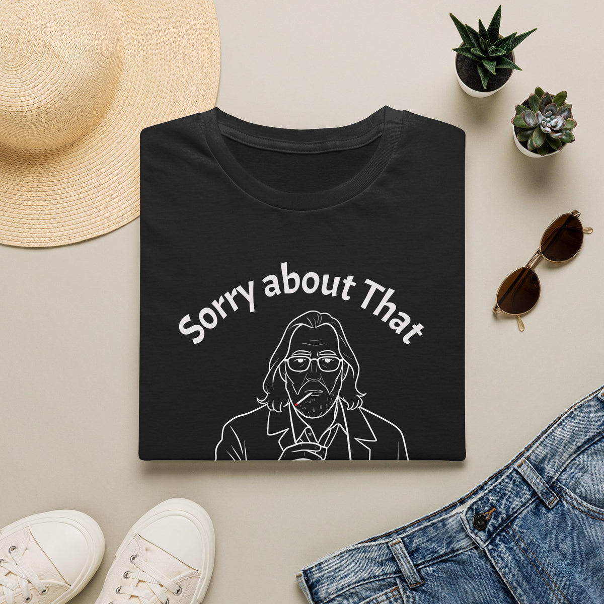 A folded black T-shirt with a white graphic print reading 'Sorry about That' above a line drawing of a man, styled in a flat lay with a straw hat, small potted succulents, sunglasses, white sneakers, and blue jeans on a light beige background.