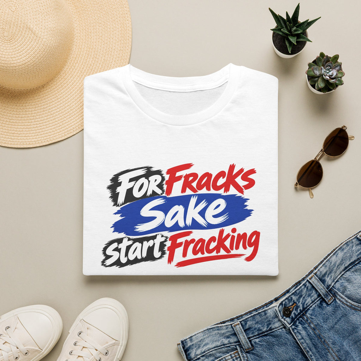 A folded white graphic T-shirt is displayed in a styled flat lay on a light beige surface. The shirt features the slogan 'For Fracks Sake Start Fracking' in red, white, and blue brushstroke-style lettering.