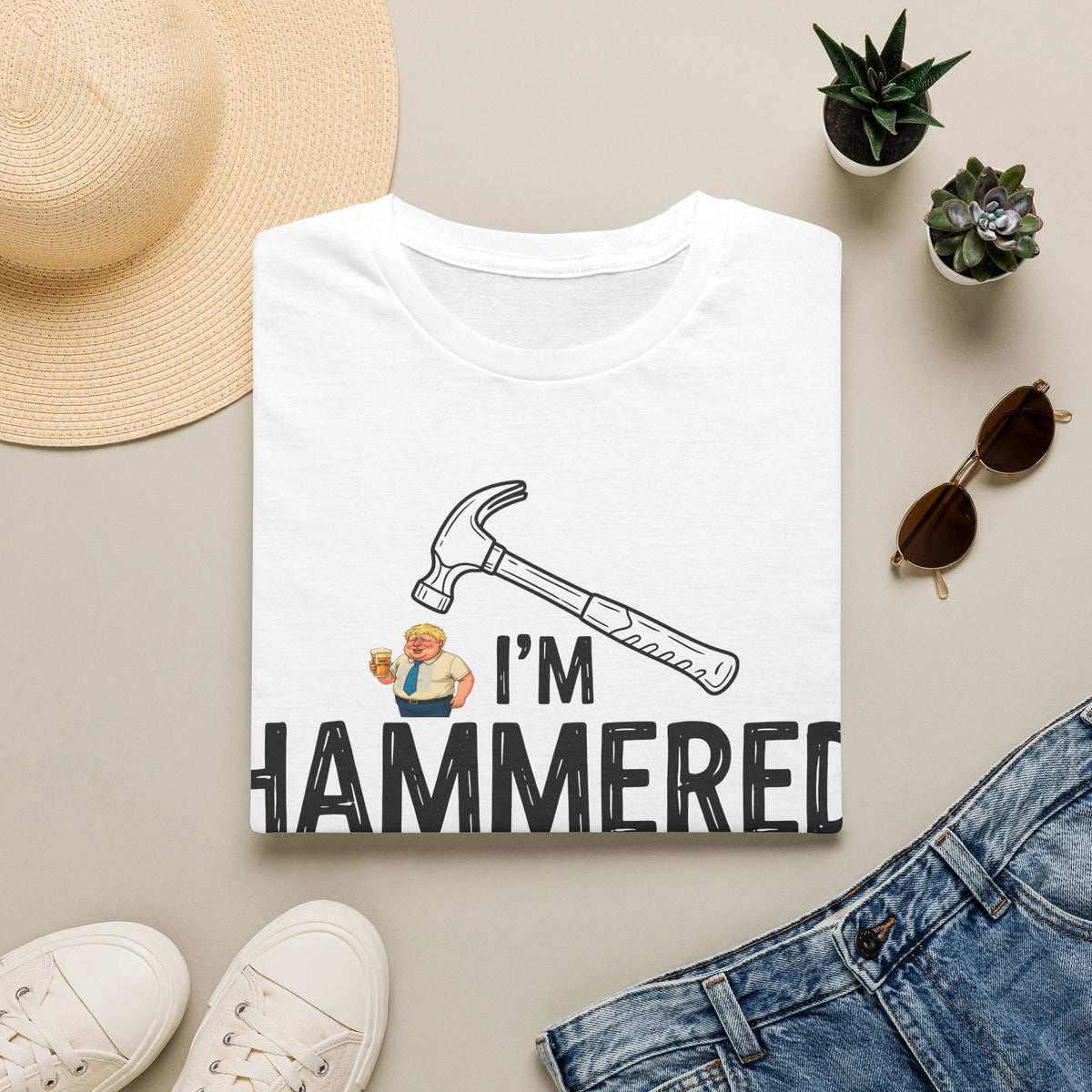 A folded white graphic T-shirt is displayed in a styled flat lay on a light beige surface. The shirt features a hammer illustration, a small cartoon figure, and the text 'I'M HAMMERED' in bold black letters.