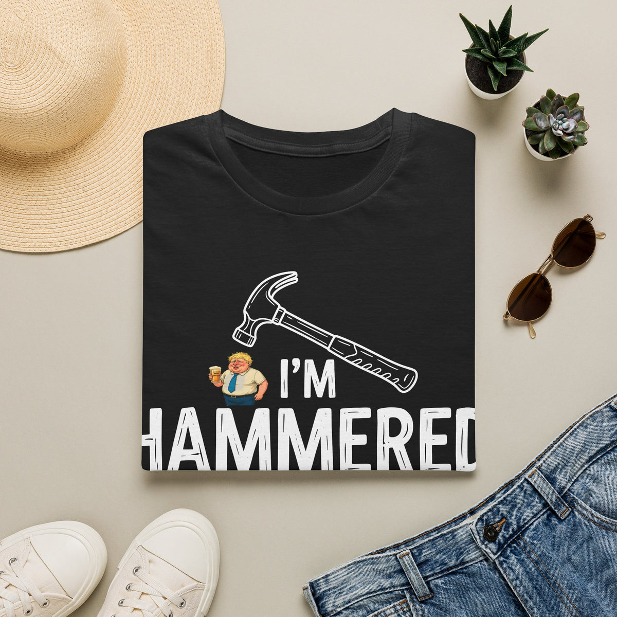 A black graphic T-shirt laid flat on a light beige surface, surrounded by a straw hat, small potted succulents, sunglasses, white sneakers, and blue jeans. The shirt graphic shows a hammer above the text 'I'M HAMMERED' with a small cartoon figure.