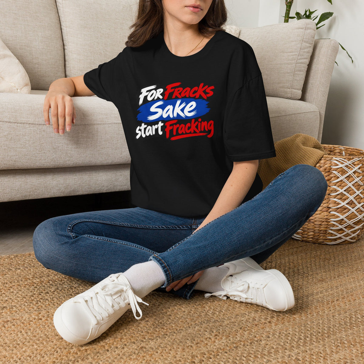 A woman sitting cross-legged on a woven rug in front of a beige sofa, wearing a black graphic T-shirt with the slogan 'For Fracks Sake Start Tracking' in white, blue, and red lettering, paired with blue jeans and white sneakers.