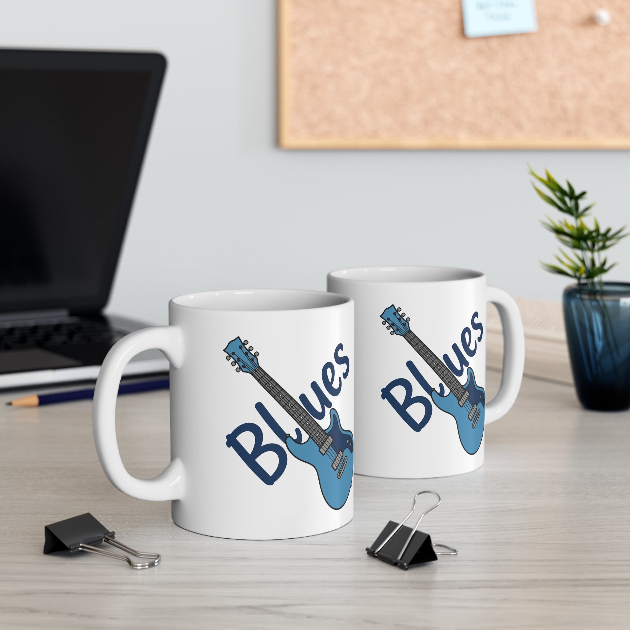 Two white mugs with blue guitars and 'Blues' text on a desk with office items.