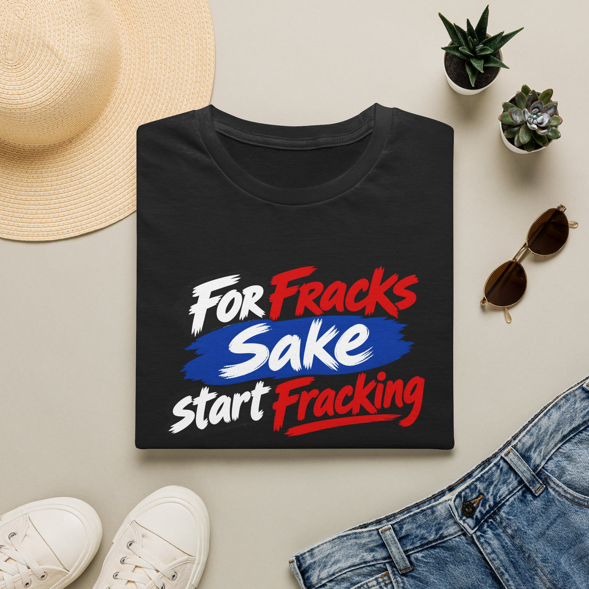 A folded black T-shirt with the slogan 'For Fracks Sake start Fracking' printed on the front in white, red, and blue, styled in a flat lay with a straw hat, white sneakers, blue jeans, sunglasses, and small potted succulents on a light beige background.
