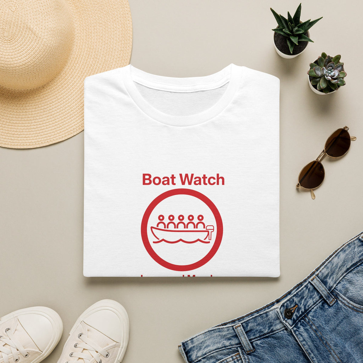 White t-shirt with a dog illustration and 'boat watch text, surrounded by a hat, sunglasses, shoes, and plants on a beige surface.