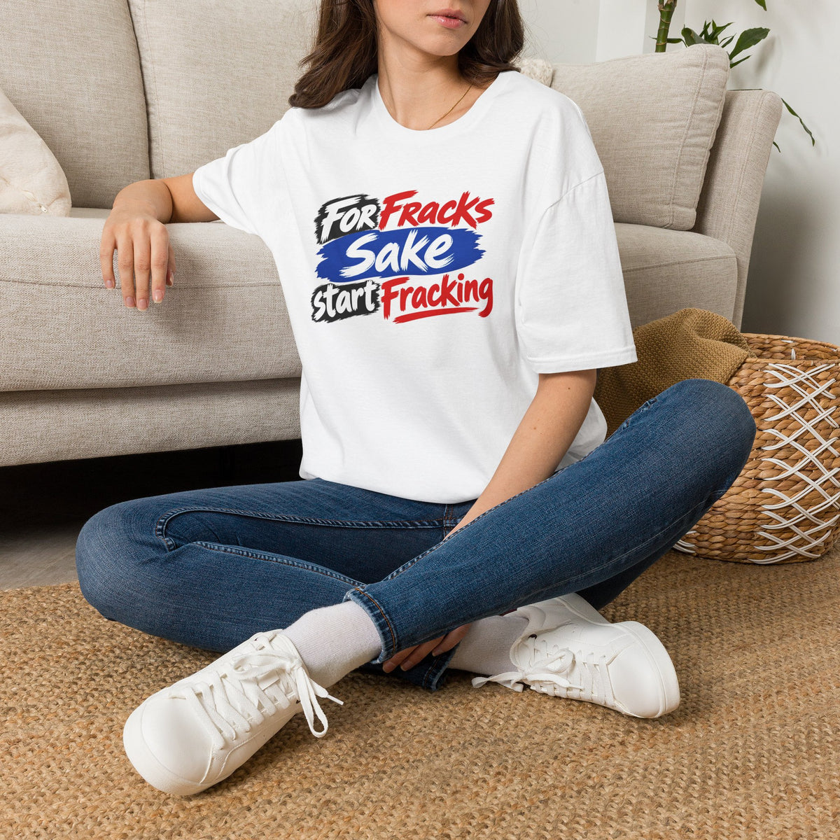 A woman sitting cross-legged on a woven rug in front of a beige sofa, wearing a white graphic T-shirt with the text 'for fraks sake start fracking, blue jeans, and white sneakers.