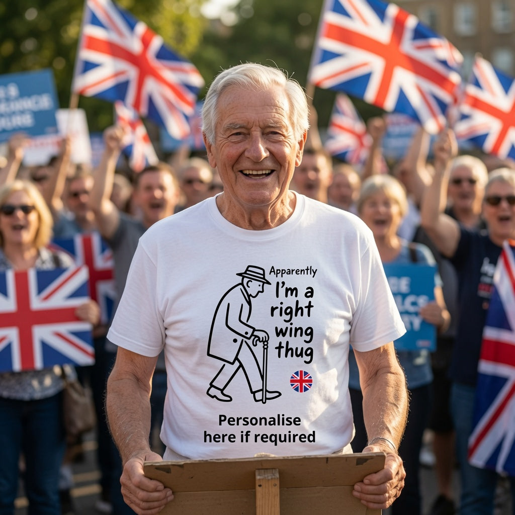 An older man smiling in a crowd, wearing a white graphic T-shirt with a black line drawing of a winged figure and the text 'Apparently I'm a right wing thug' plus a small Union Jack flag and 'Personalise here if required'. People behind him hold Union Jack flags and signs.