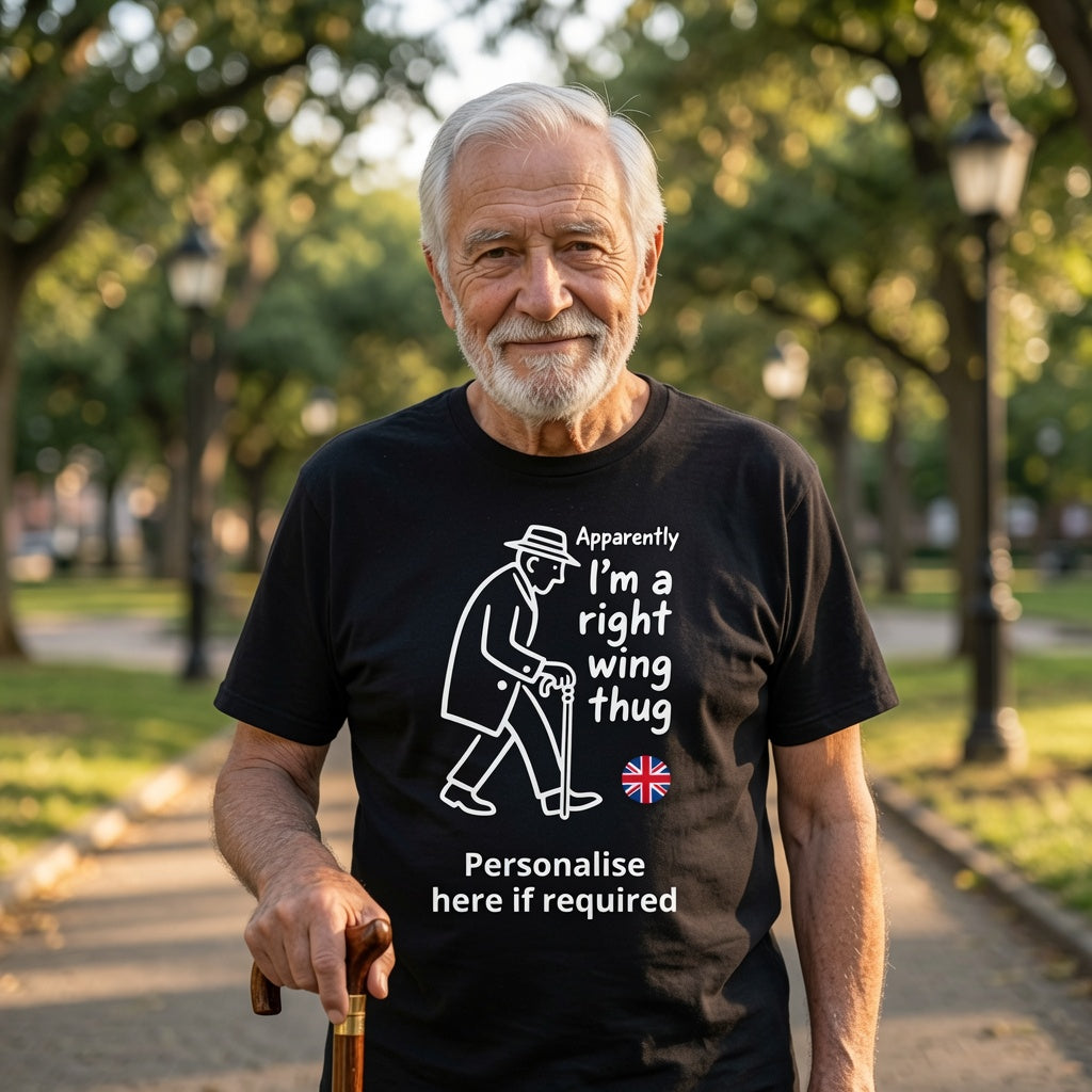 An older man with white hair and a beard stands on a park path wearing a black graphic T-shirt. The shirt has a white line drawing of an elderly man with a cane and the text 'Apparently I'm a right wing thug' along with a small Union Jack graphic and the words 'Personalise here if required'.