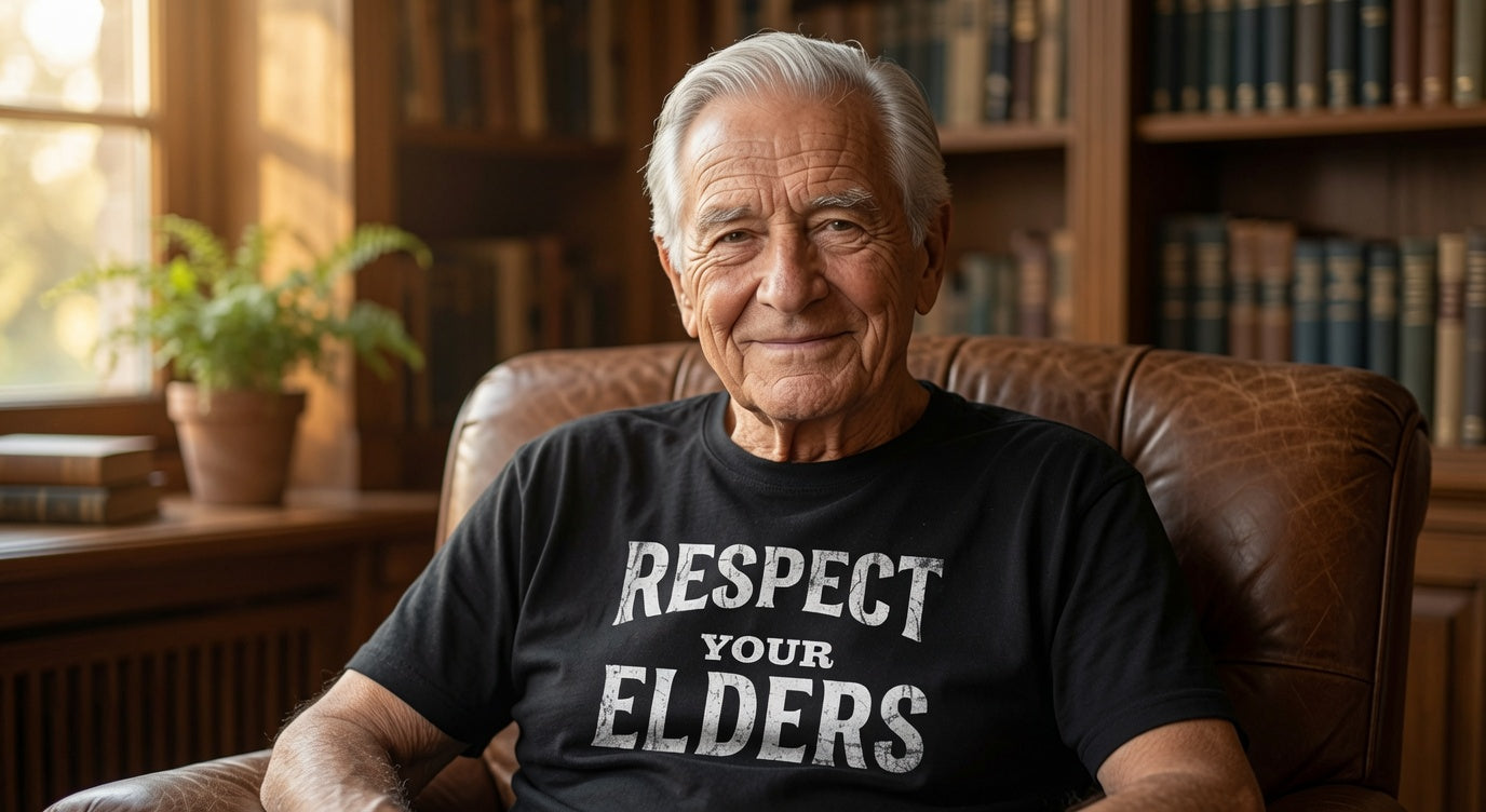 An older man sitting in a brown leather armchair in a library or study, wearing a black t-shirt with the text 'RESPECT YOUR ELDERS' printed in white. The background features wooden bookshelves filled with books.