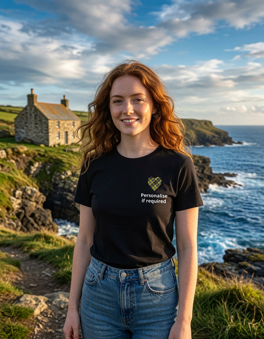 A smiling woman with red hair stands outdoors by the ocean wearing a black graphic T-shirt and blue jeans. The shirt has a Cornish tartan green heart-shaped logo and white text reading 'Personalise if required.
