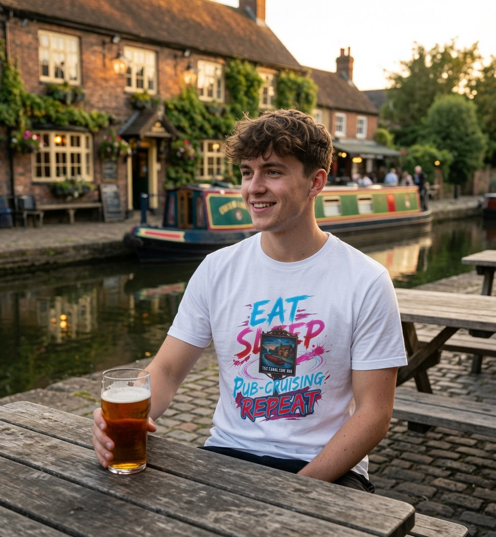 A young man sitting at a wooden pub table outdoors by a canal, wearing a white graphic t-shirt with the text 'EAT SLEEP PUB-cruising REPEAT' and holding a pint of beer.