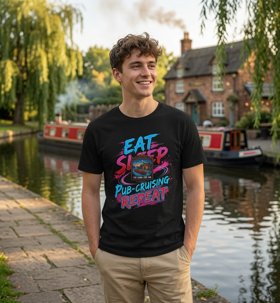 A smiling young man stands by a canal with a brick pub and boats in the background, wearing a black graphic T-shirt with colorful text reading 'EAT SLEEP PUB-CRUISING REPEAT' and khaki pants.