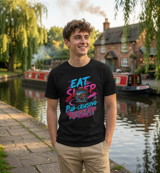 A smiling young man stands by a canal with a brick pub and boats in the background, wearing a black graphic T-shirt with colorful text reading 'EAT SLEEP PUB-CRUISING REPEAT' and khaki pants.