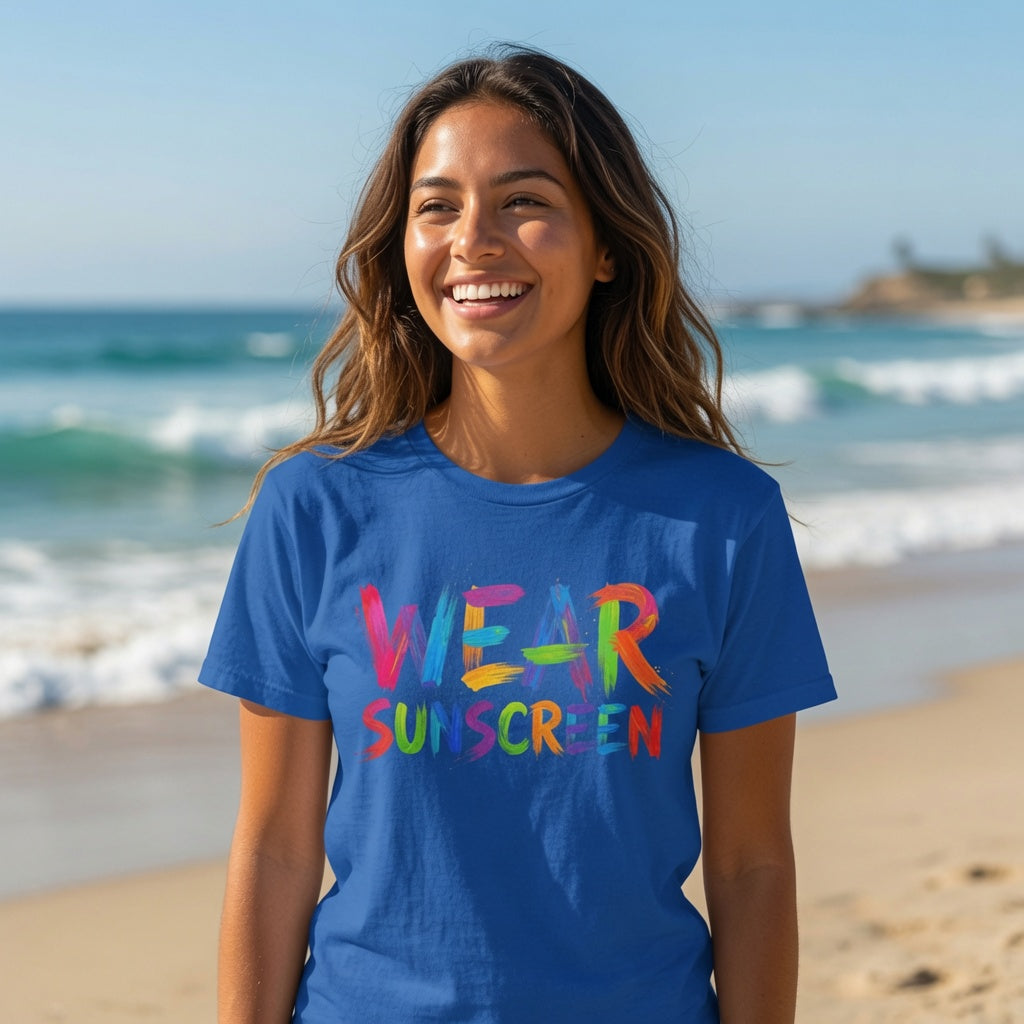 A woman smiling on a sunny beach wearing a royal blue t-shirt with 'WEAR SUNSCREEN' printed in a colorful rainbow font.