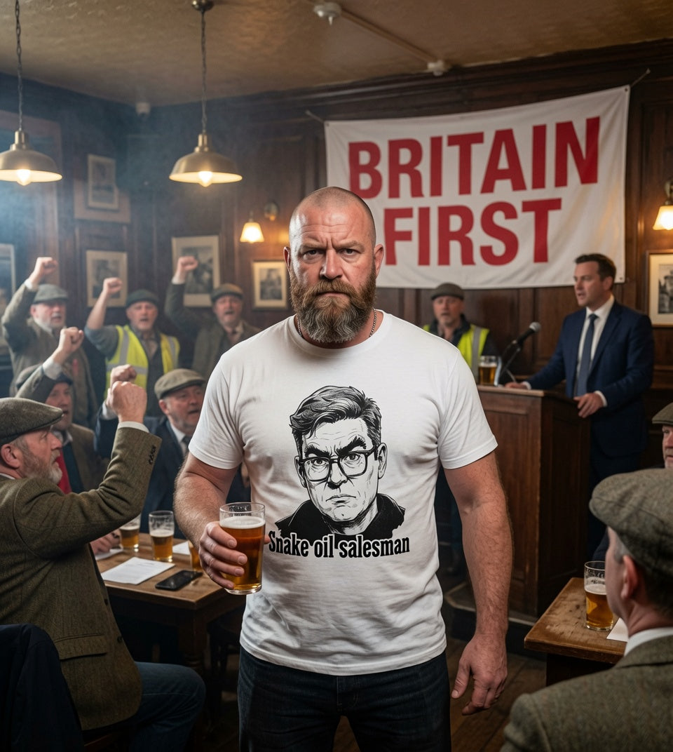 A man in a pub wearing a white graphic t-shirt with a black-and-white portrait of Andy Burnham and the text 'Snake oil salesmen' and 'BRITAIN FIRST' hanging in the background.