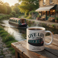 White mug with 'Live Life at 4mph' text and steam on a wooden table by a canal with a boat and building in the background.
